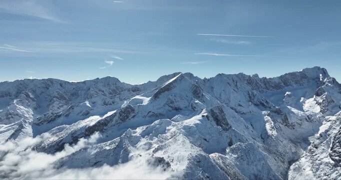 Aerial view of snow-capped mountains piercing through the clouds under a clear blue sky, creating a striking contrast in Garmisch-Partenkirchen, Bavaria, Germany.