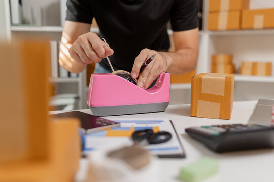 Small business owner sealing parcel with tape dispenser while preparing packages for shipment, organizing delivery orders and packaging process, representing ecommerce and logistics workflow.