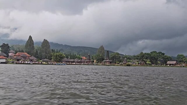 POV video from a boat showing Pura Ulun Danu Beratan temple on the lake, featuring multi-tiered pagoda roofs, water surface, surrounding hills, and light mist.
