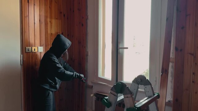 Interior renovation scene of a worker using a crowbar to remove wooden wall paneling near a window. DIY home improvement concept with natural daylight and construction tools.