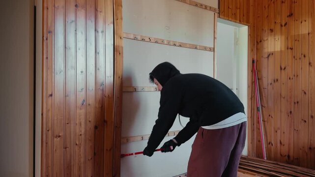 Worker using crowbar to dismantle wooden wall panels inside a house. Interior renovation and demolition process with construction tools and debris.