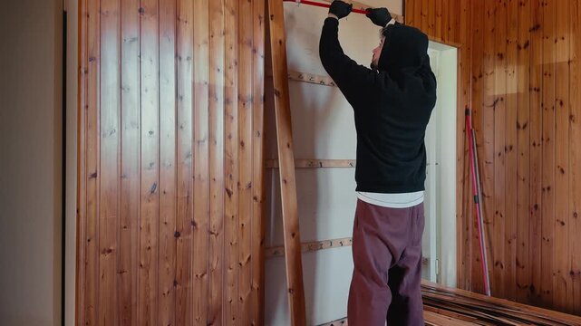Man using a crowbar to pry off wooden wall panels in a house. Interior demolition and renovation process with visible structure and construction work in progress.