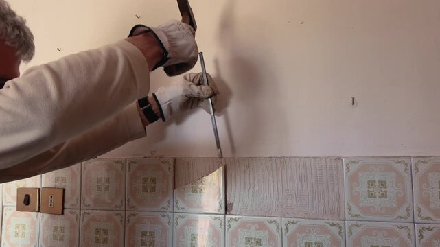 Close-up of a worker using a chisel to remove old ceramic wall tiles during interior renovation. Damaged plaster and exposed wall surface visible in a home demolition scene.