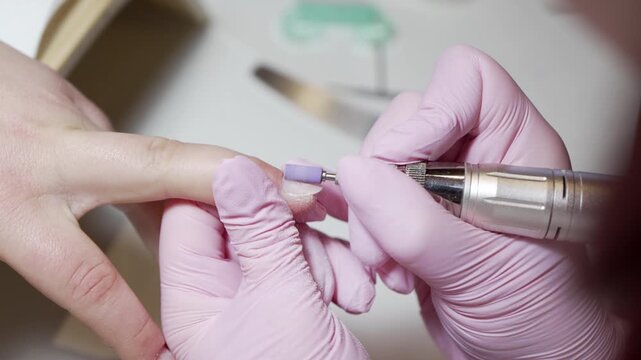 Close up of a nail technician in pink gloves using an electric drill file to remove gel polish and clean the client's cuticles