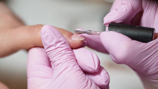 Professional beautician in pink gloves applying a transparent base coat polish on a client's fingernail. Nail care and beauty treatment