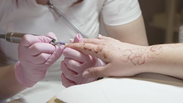Professional manicurist in pink gloves using an electric drill to file and shape a client's fingernails and remove cuticles in a beauty salon