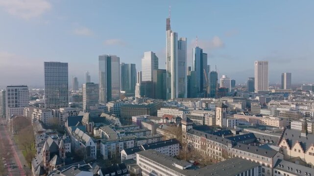 Aerial view of the Frankfurt skyline with the prominent Commerzbank Tower at its center, surrounded by other skyscrapers and the city center under a blue, slightly cloudy sky.