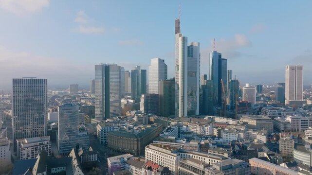 Aerial view of the Frankfurt skyline with the prominent Commerzbank Tower at its center, surrounded by other skyscrapers and the city center under a blue, slightly cloudy sky.