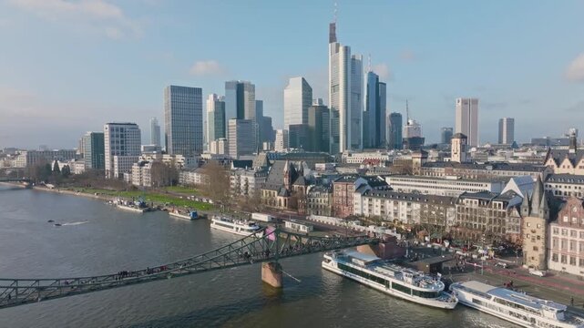 Aerial view of the Eiserner Steg footbridge in Frankfurt with the prominent skyscraper skyline in the background. Excursion boats are docked at the Main riverbank under a clear sky.