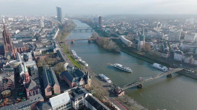 Aerial view of Frankfurt: Main river with bridges and boats, Frankfurt Cathedral and old town on the left, modern skyline in the background under a clear sky.