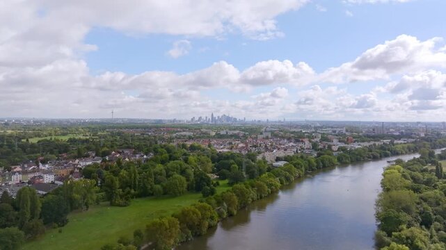 A wide aerial view of the Main River winding through a lush green landscape. In the background, the prominent skyline of Frankfurt am Main is visible under a slightly cloudy summer sky.