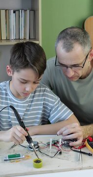 Boy and adult assembling electronic circuit using soldering iron and tools at home workspace. Father and son have a shared hobby. Concept of DIY electronics, hobby engineering, and maker. Vertical.