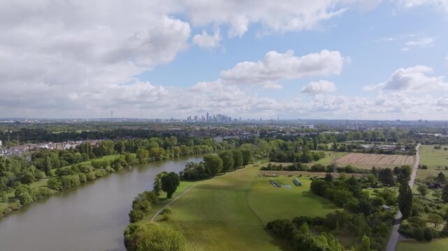 A wide aerial view of the Main River winding through a lush green landscape. In the background, the prominent skyline of Frankfurt am Main is visible under a slightly cloudy summer sky.