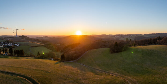 Aerial view of the sun setting behind wind turbines casting long shadows over rolling green fields, Hellenthal, Germany.