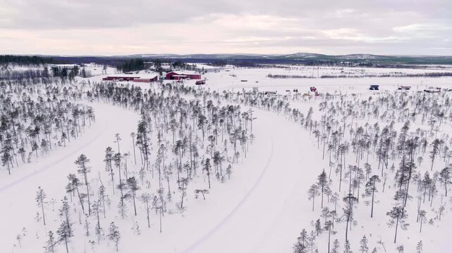 Aerial view of a wintry plain in Finland. It shows snow-covered fields, scattered conifers, and a small group of red wooden houses under a vast, overcast sky.