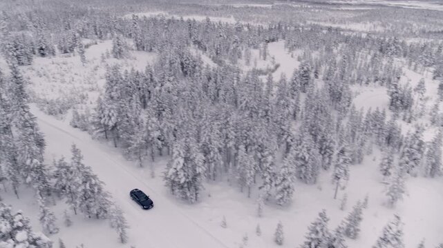 Aerial view of a wintry plain in Finland. It shows snow-covered fields, scattered conifers, and a small group of red wooden houses under a vast, overcast sky.