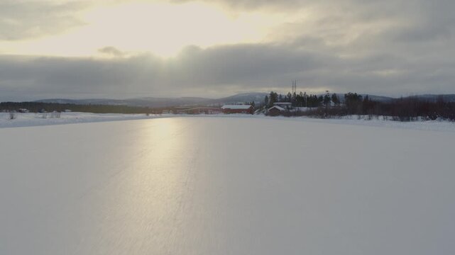 A vast, smooth frozen ice surface in Finland under a cloudy winter sky. In the background, typical red wooden houses and dark coniferous forests stand before a range of gentle hills.