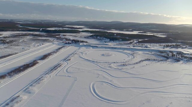 Broad aerial view of a snowy testing ground in Finland under a clear winter sky. The image shows various circular and curved driving tracks in the snow, as well as long, straight testing lanes embedde