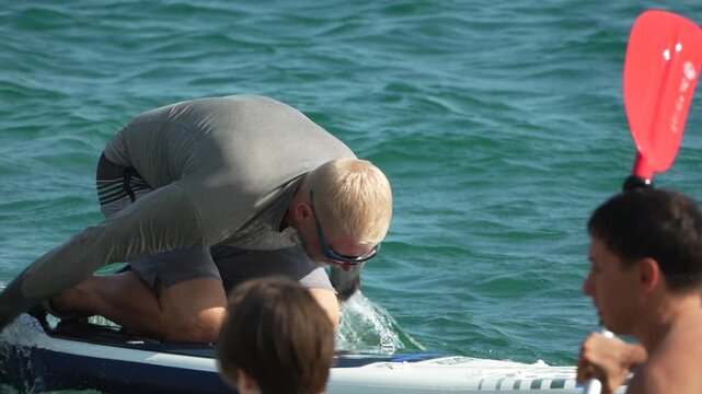 Paddleboard ocean man kneeling on a board in the turquoise sea and adjusting sunglasses during a summer vacation day of water sports