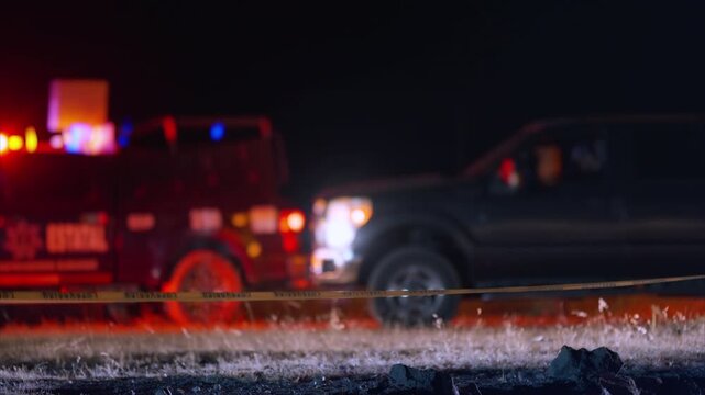 Yellow caution tape cordons off nighttime emergency scene with blurred emergency vehicles and flashing red and blue lights in background creating dramatic tense atmosphere on dark road.