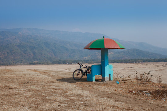 Sunamganj, Bangladesh - 26 January 2019: View of a solitary blue resting spot with a vibrant red and green umbrella-shaped roof stands juxtaposed against the vast, arid landscape and distant hills.