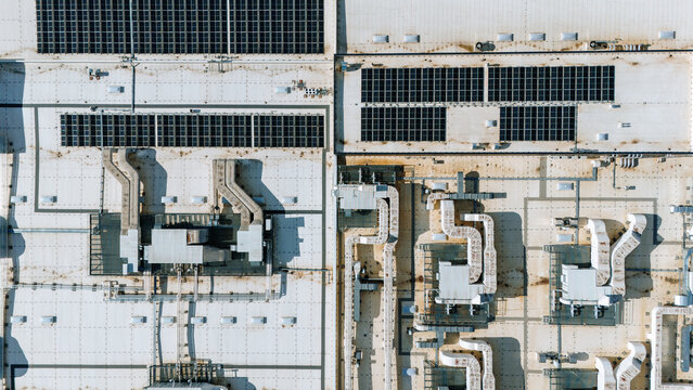 Aerial view of rows of solar panels glinting under the sun atop vast industrial rooftops, casting stark shadows across the utilitarian landscape, Becej, Vojvodina, Serbia.