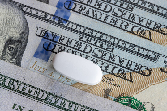 A white pharmaceutical tablet resting atop U.S. hundred-dollar banknotes. The recognizable currency design, featuring "The United States of America"