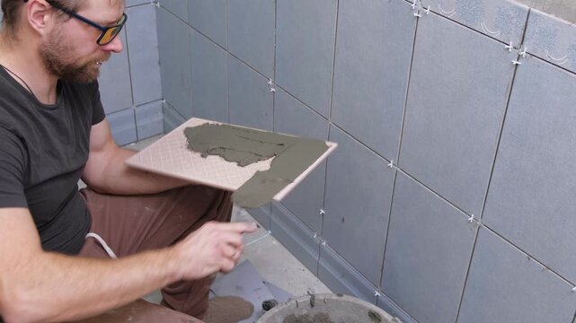Tile adhesive spreading. Tiling mortar application. Worker smoothing adhesive. Workman smoothing grout on ceramic tiles. Bathroom renovation. Man using notched trowel, cement spread across tile back