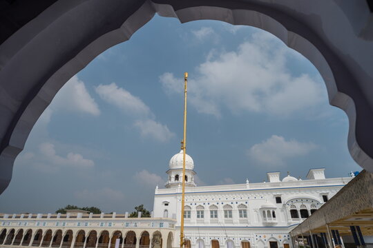 View of the golden spire rising boldly above the white Gurdwara, framed by an arch against a blue sky, a serene vista, Nankana Sahib, Punjab, Pakistan.