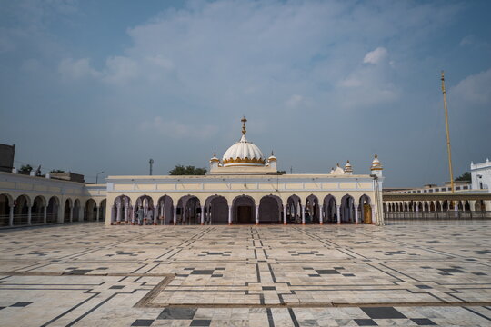 View of a pristine white structure with arched entrances and a gleaming dome stands on a patterned courtyard under a vast sky, Nankana Sahib, Punjab, Pakistan.