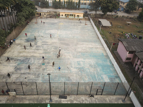 Aerial view of people scattered across a weathered sports court, framed by buildings and trees in a tightly packed urban setting, Lagos, Lagos, Nigeria.