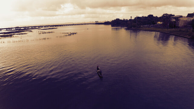 Aerial view of a lone boat glides across the water towards the distant bridge in Oworonshoki, under a sky of muted tones, Oworonshoki, Lagos, Nigeria.