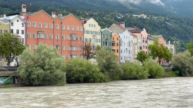 Static shot on overcast day showing iconic colorful Inn riverfront buildings in Innsbruck with misty Nordkette Alps behind.