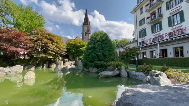 Static shot, partly cloudy sunny day, Japanese garden koi pond with church steeple and Hotel Interlaken, serene Swiss atmosphere.