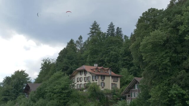 Static shot, overcast dramatic clouds, paragliders soaring above traditional Swiss chalets and dense forest hillside in Interlaken, calm grey mood.