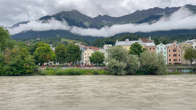Static shot of turbulent muddy Inn River with colorful historic buildings, lush trees, and cloud-shrouded Nordkette Alps in Innsbruck, Austria.