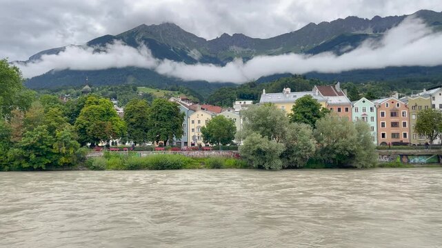 Static shot of turbulent Inn River with colorful historic buildings, Nordkette Alps partially shrouded in low clouds, Innsbruck, Austria.