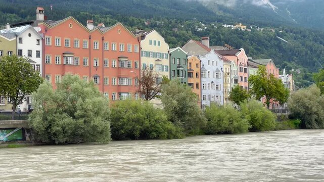 Static shot on overcast day showing vibrant historic colored facades along Inn River with Nordkette Alps behind, Innsbruck, Austria.