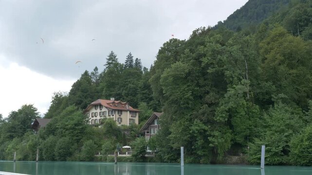 Static shot on overcast day, multiple paragliders soaring above turquoise Aare River, alpine houses, lush forested hills, Interlaken Switzerland.
