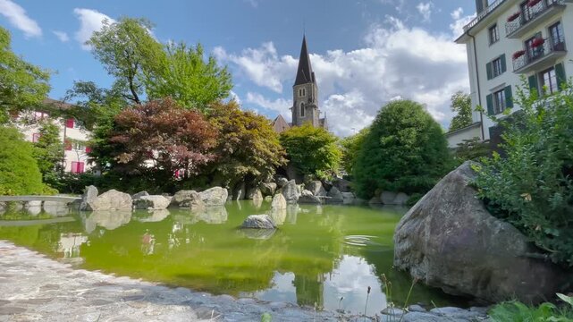 Static shot of a serene Japanese garden pond with rocks, green algae water, and Gothic church spire in Interlaken, Switzerland, under partly cloudy summer sky.