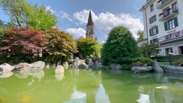 Static shot, sunny partly cloudy day, Japanese garden with green koi pond, rocks, colorful trees, church steeple and hotel, Interlaken Switzerland.