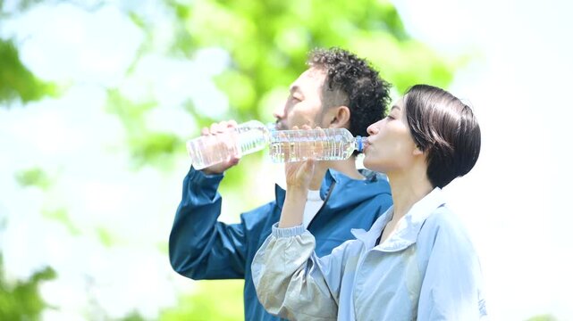 新緑の屋外でスポーツの合間に水分補給、ペットボトルの水を飲む夫婦の動画スローモーション