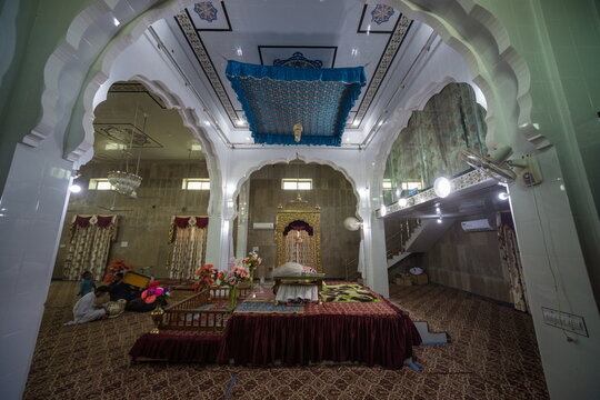 View of an elaborately decorated interior space with ornate arches, a draped canopy, and a raised platform draped in rich red fabric, Nankana Sahib, Punjab, Pakistan.