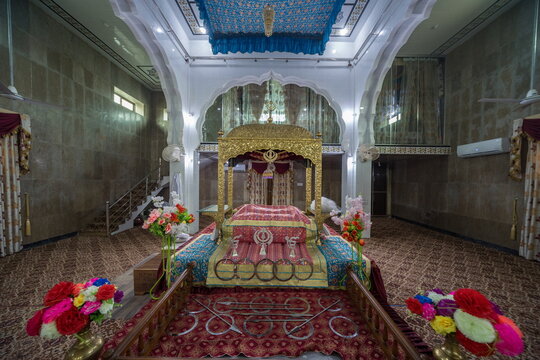 View of a golden palanquin draped in pink and gold cloth, set against marble walls and a patterned carpet, bathed in soft light, Nankana Sahib, Punjab, Pakistan.
