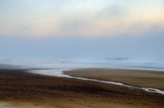 View of a stream of water flows into the sea under a layer of fog, where the sky fades in a gradient of soft blues and whites, Hossegor, France.