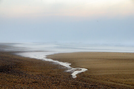 View of a sinuous stream reflecting the soft, diffused light, cutting through the tranquil beach on a misty day, Hossegor, France.