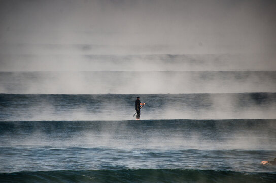 View of a lone paddleboarder cutting through the misty ocean waves, creating a stunning contrast between the dark water and ethereal fog, Hossegor, France.