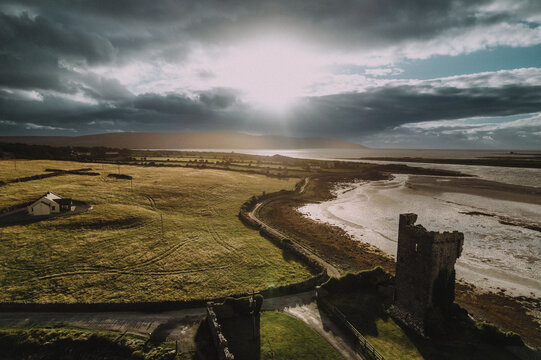 Aerial view of sunbeams piercing through dramatic clouds over green fields and the Ballyportry Castle ruins, County Clare, Ireland.