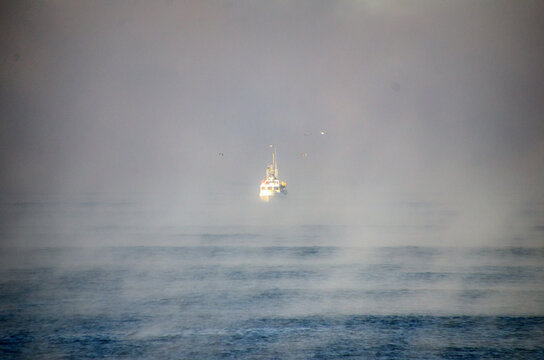 View of a solitary white boat piercing through the dense, ethereal fog blanketing the vast, blue ocean, a serene yet haunting scene, Hossegor, France.
