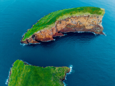 Aerial view of the craggy Ilheus das Cabras islands, with verdant green vegetation contrasting with the rugged, reddish-brown cliffs, Terceira, Azores, Portugal.
